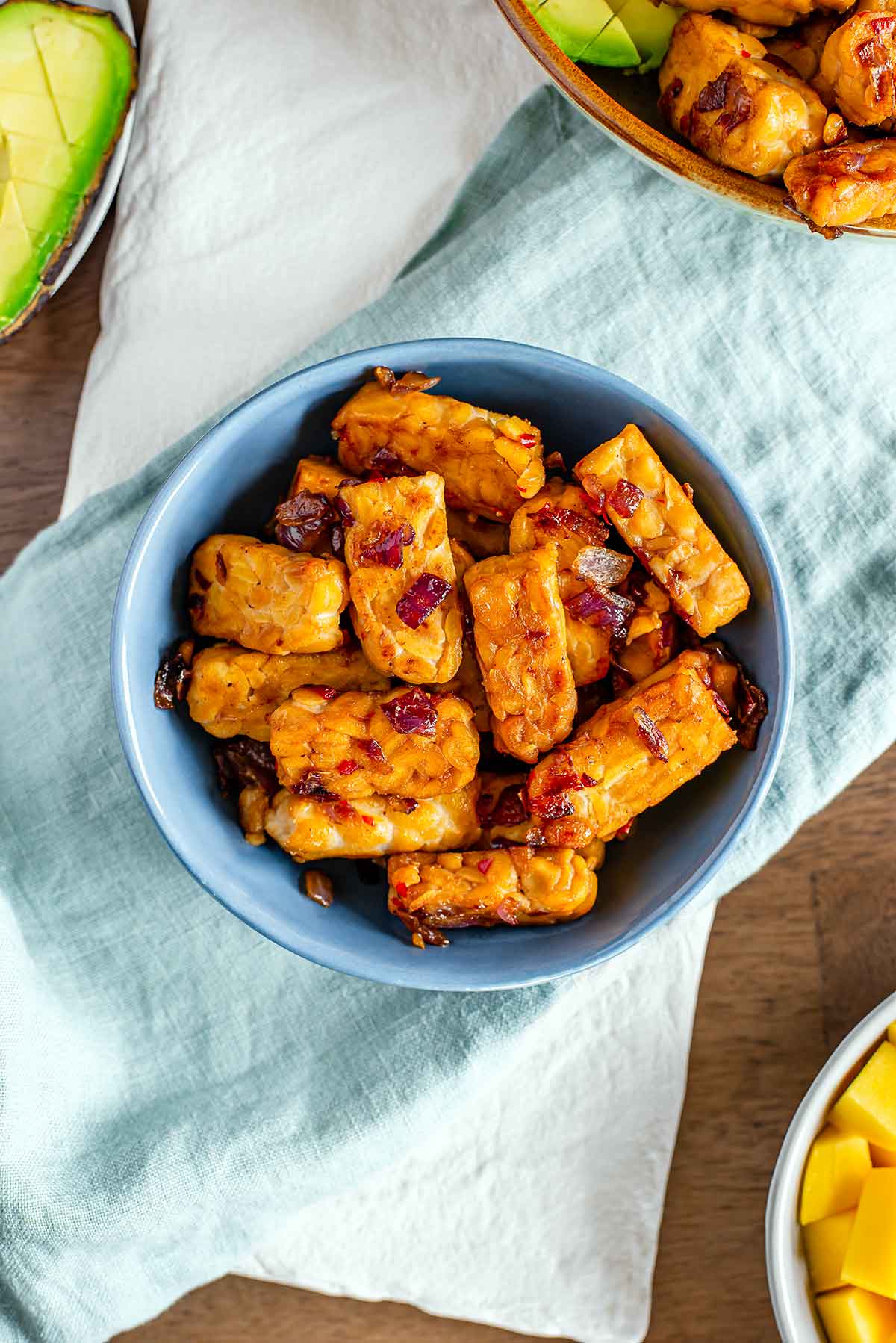 Top down view of simple stovetop tempeh in a small bowl atop a white tray. Sautéed red onion and flecks of red chili can be seen on the golden brown sweet and spicy tempeh.