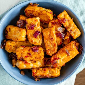 Top down view of sweet and spicy pieces of tempeh in a small bowl. Sautéed red onion and flecks of red chili can be seen on the golden brown tempeh.