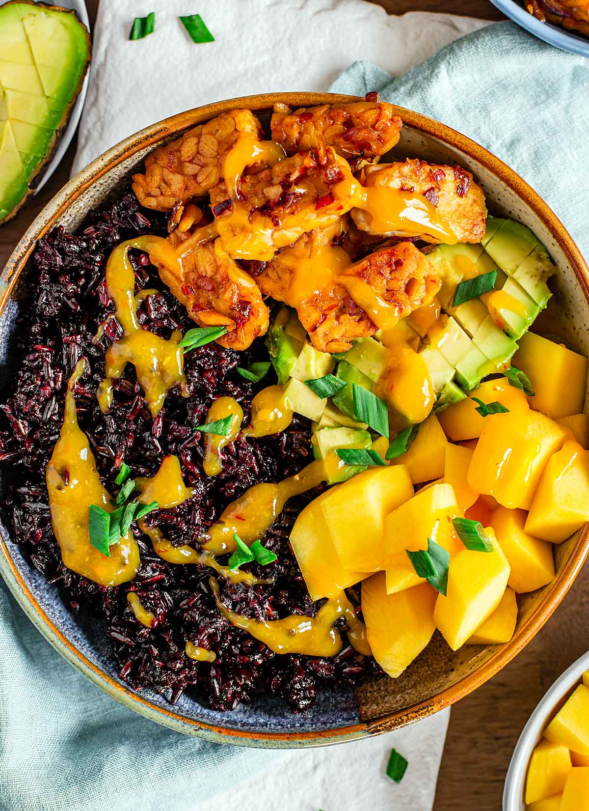 Top down view of simple stovetop tempeh served as part of a summery rice bowl. Black rice, cubed mango, and diced avocado surround the tempeh. The bowl is drizzled with a bright yellow mango chili sauce.