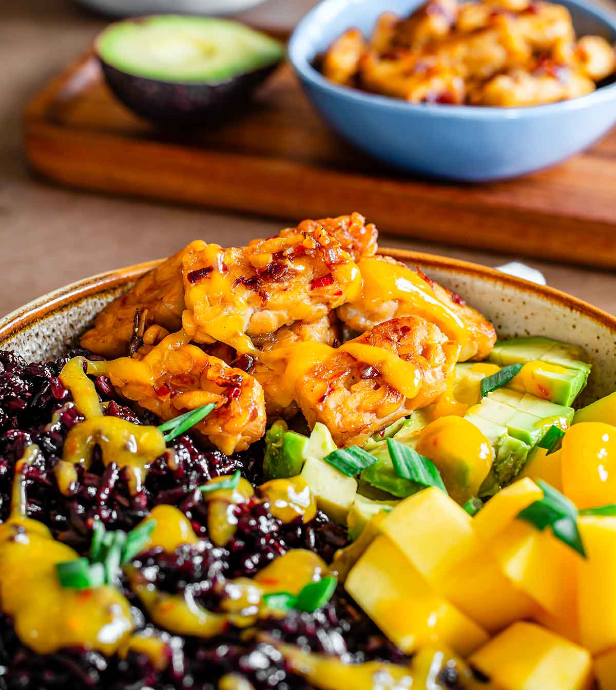 Side view close up of tempeh on a summery black rice bowl with mango and avocado. The tempeh is golden brown and drizzled with a bright yellow mango chili sauce.