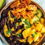 Close up view of simple stovetop tempeh served as part of a summery rice bowl. Black rice, cubed mango, and diced avocado surround the tempeh. The bowl is drizzled with a bright yellow mango chili sauce.