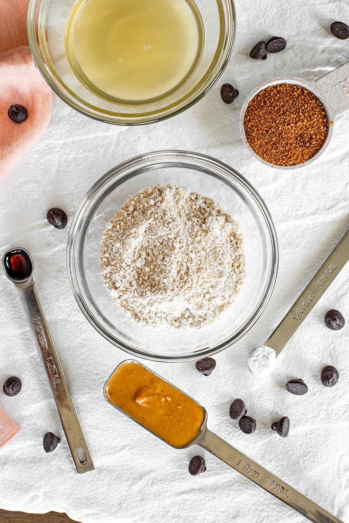 Top down view of ingredients on a white tray speckled with chocolate chips. A small bowl of oat flour is surrounded by a bowl of aquafaba and measuring spoons of coconut sugar, peanut butter, vanilla extract, and baking soda.