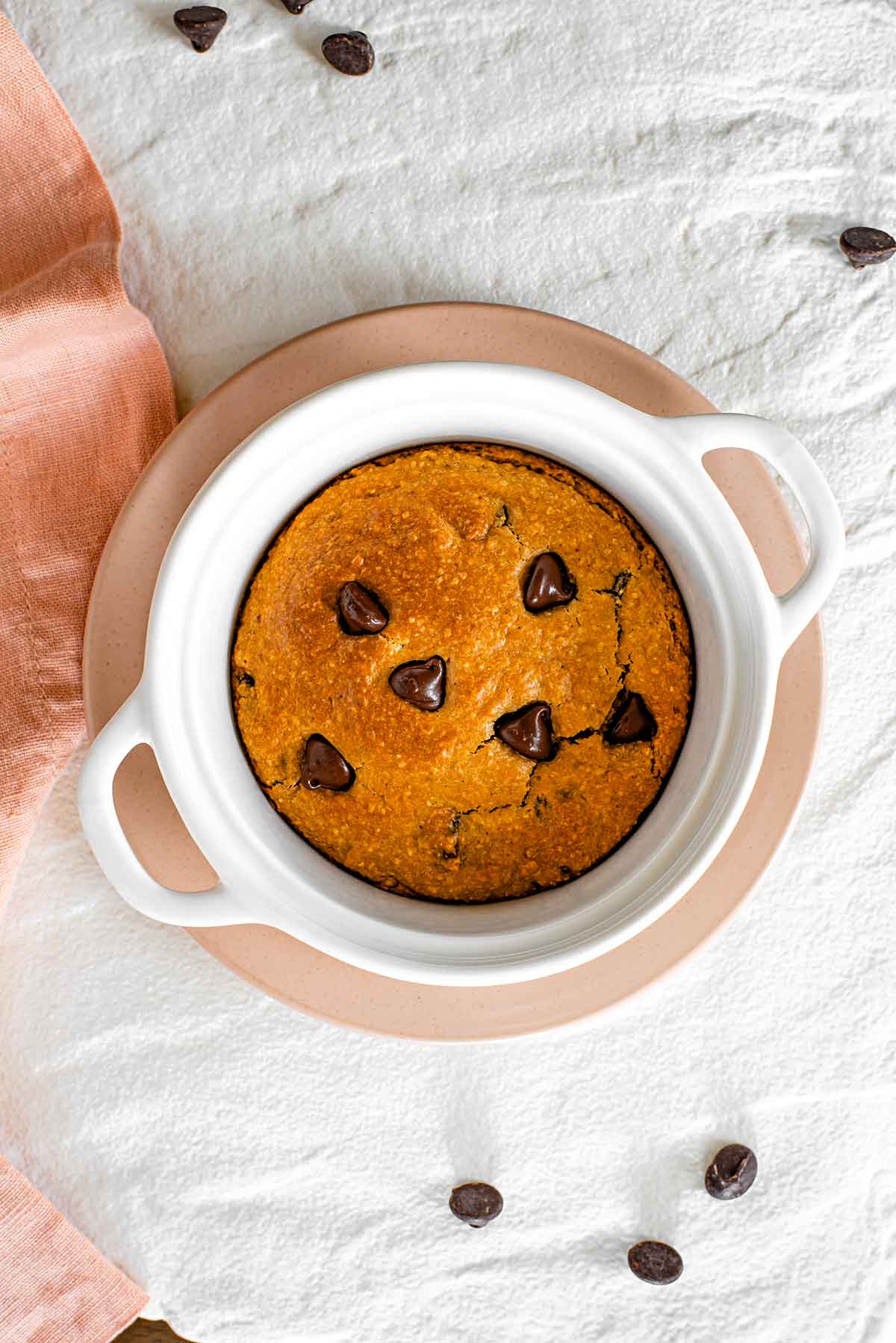 Top down view of a vegan deep dish cookie in a mini casserole dish on top of a white tray. The cookie is golden brown, speckled with chocolate chips, and has a crispy edge that has pulled away from the baking dish.