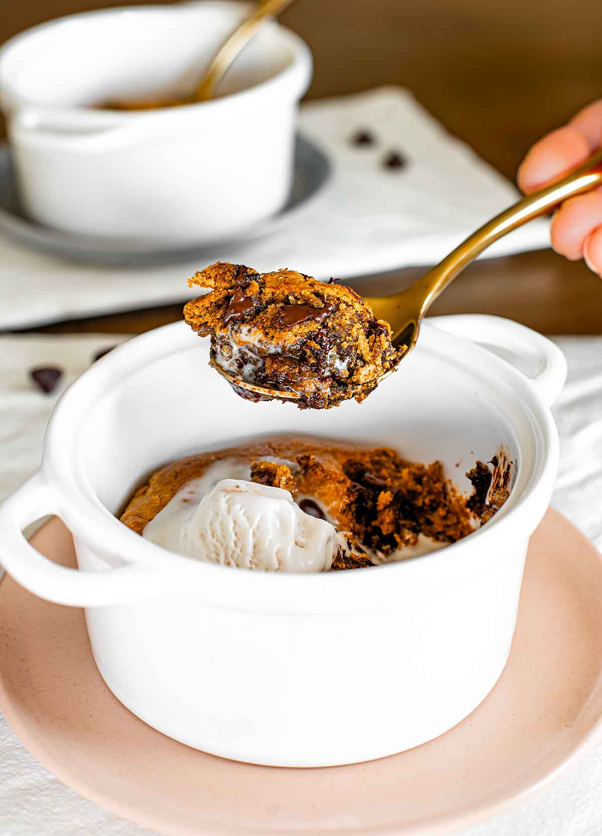 A spoon holds up a scoop of a warm and gooey vegan deep dish cookie. The cookie is crispy on the outside, soft on the inside, and full of melted chocolate. A dollop of vanilla ice cream melts on top of the remaining cookie in the baking dish.