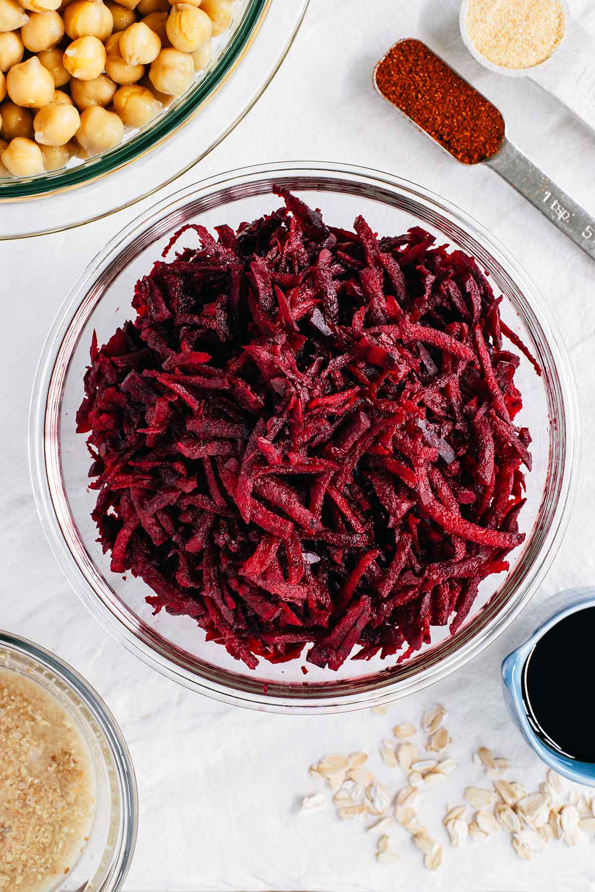 Top down view of ingredients on a white tray. Shredded beetroot fills a glass bowl in the centre with chickpeas, a flax egg, and seasonings scattered around.