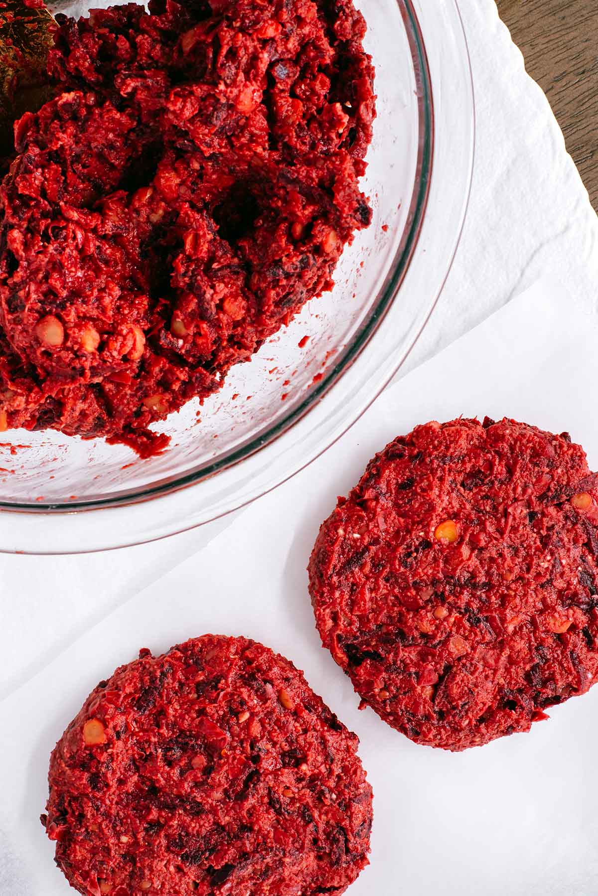 Top dow view of the mixed batter for vegan beet burgers in a glass bowl. The mixture is moist but not overly wet. Beside the bowl lay two formed beet burger patties.