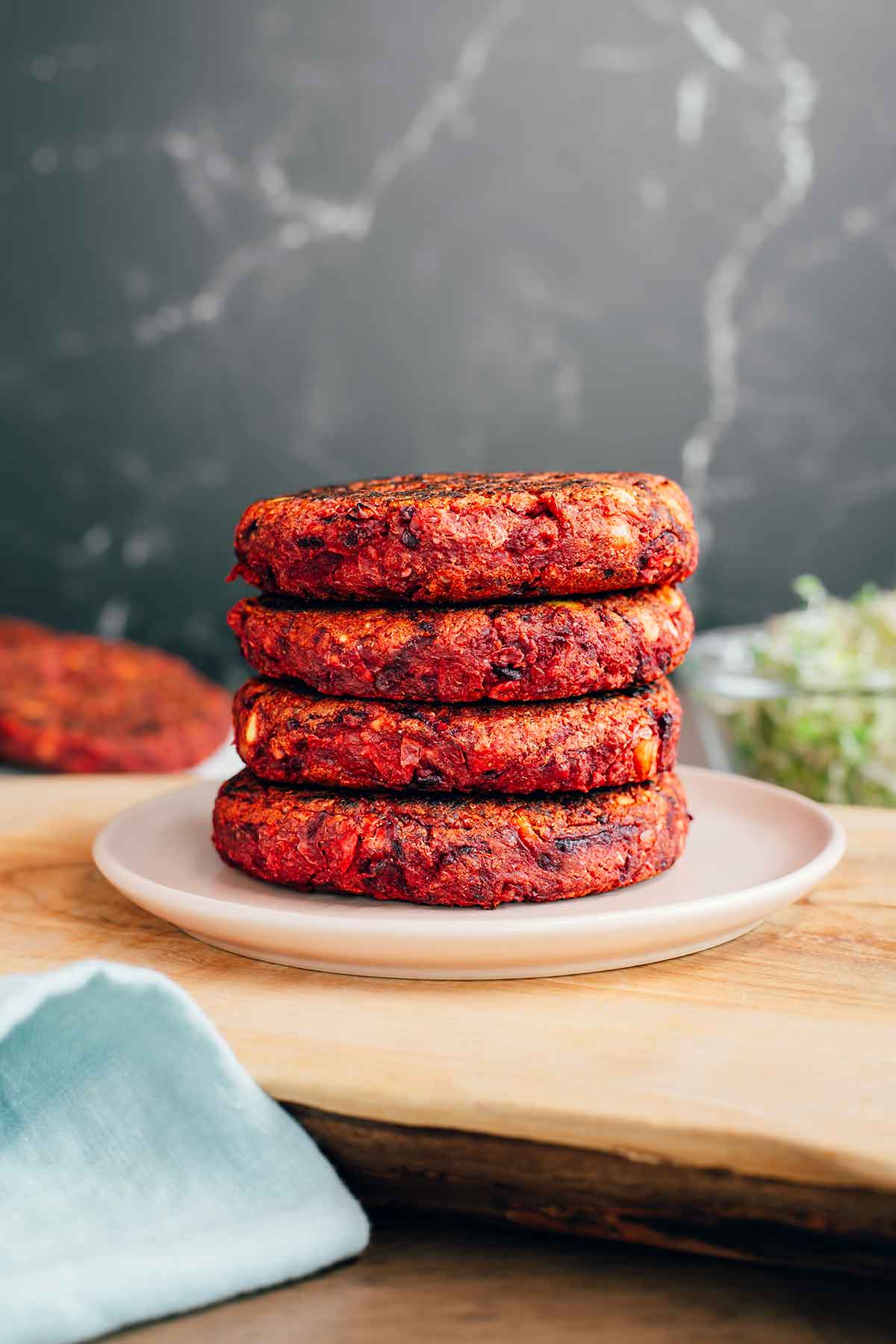 Side view of four vegan beet burger patties stacked on a small plate atop a wooden tray. The burgers are thick, sturdy and a beautiful deep red colour. 