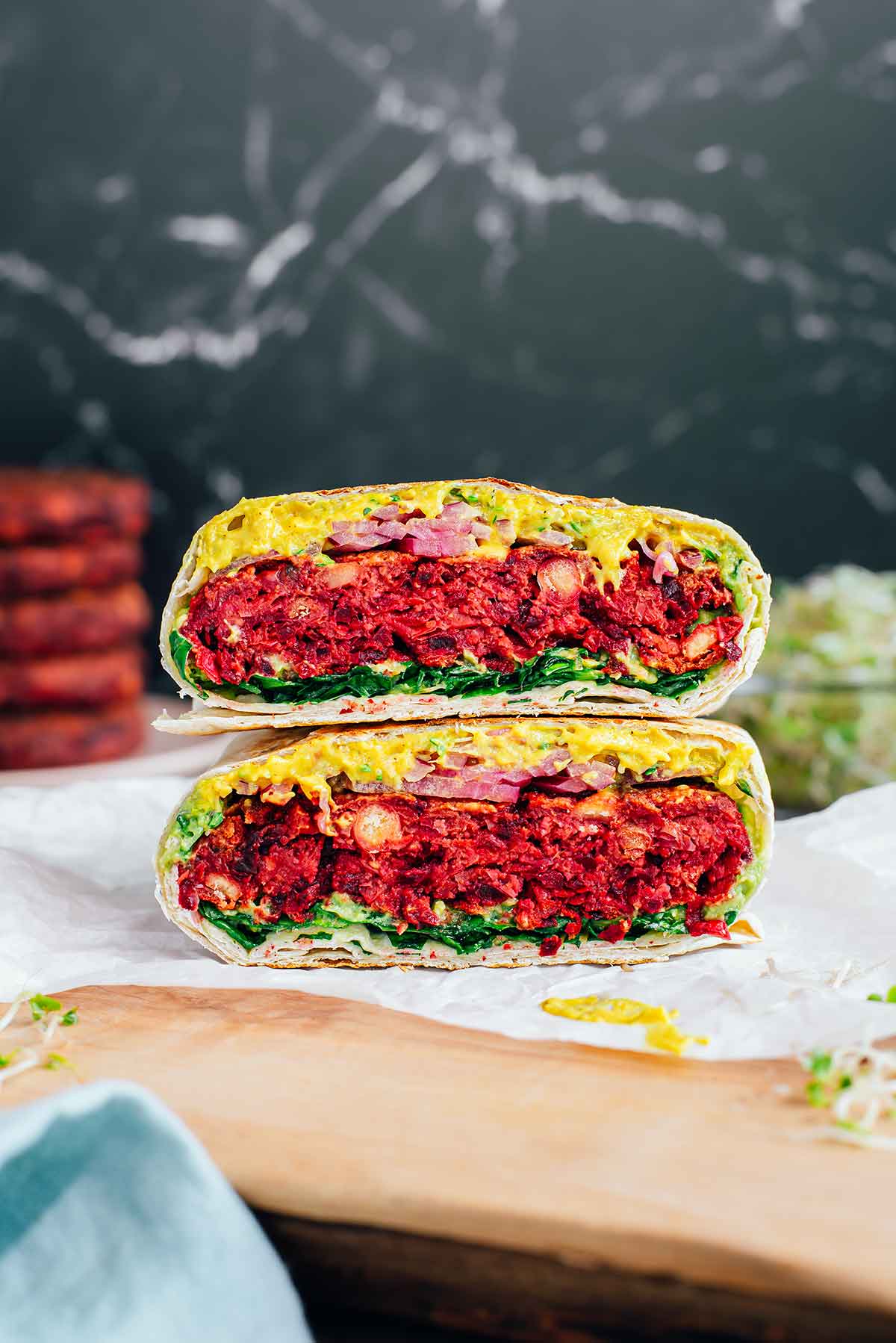 Side view of two halves of a beet burgeritto stacked on top of one another. A thick and juicy red beet burger patty is surrounded by a vegan cheese sauce, sprouts, pickled red onion, mashed avocado, and chopped greens. Everything is encased in a toasted burrito shell instead of a traditional burger bun. 