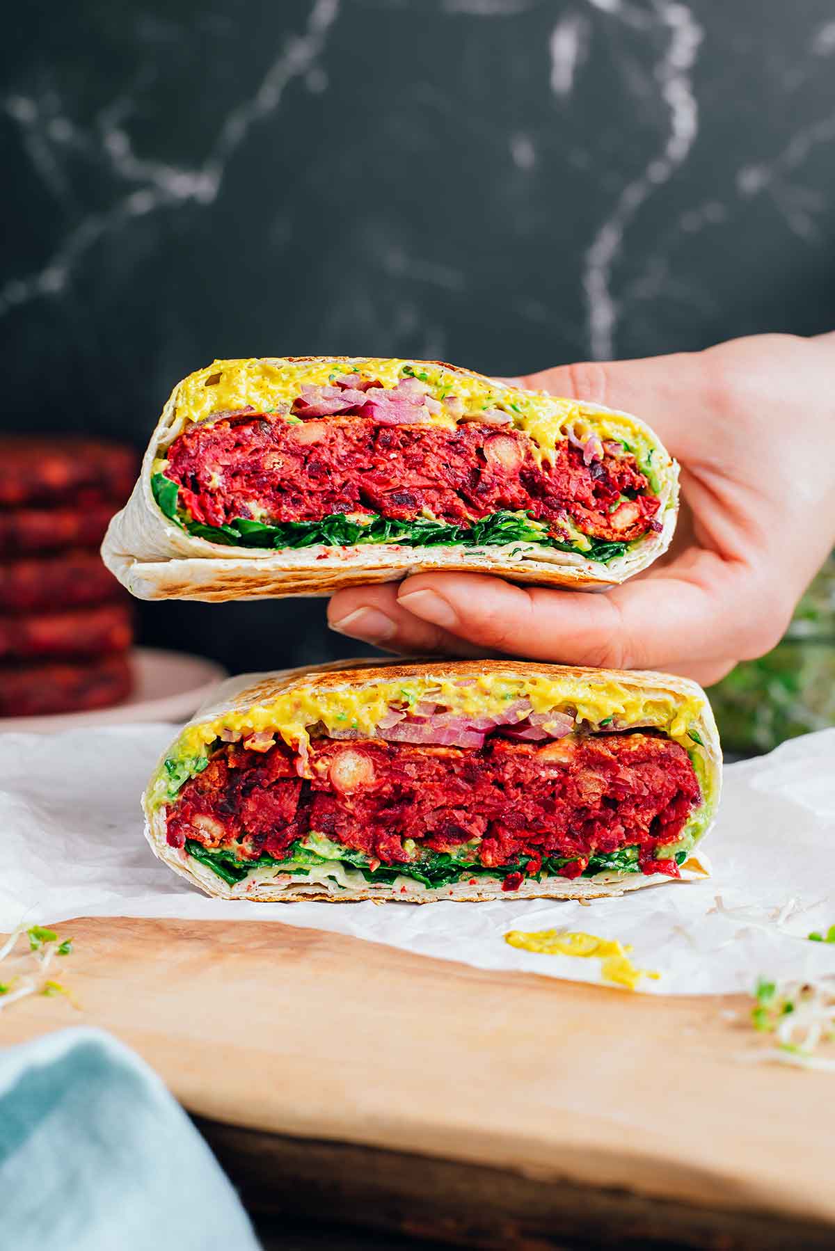Side view of a hand holding up one half of a beet burgeritto. The thick burger bun fills the seared tortilla shell and is covered in vegan queso, sprouts, mashed avocado, pickled red onion, and chopped spinach.