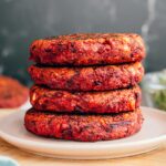 Side view of four vegan beet burger patties stacked on a small plate. The burgers are thick, sturdy and a beautiful deep red colour.