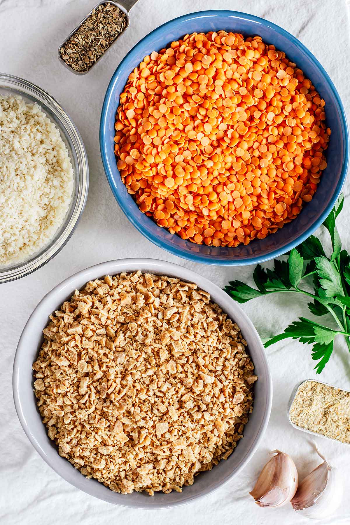 Top down view of ingredients on a white tray. A small bowl of dehydrated TVP rests beside a bowl of split red lentils. Breadcrumbs, some seasonings and fresh parsley surround.