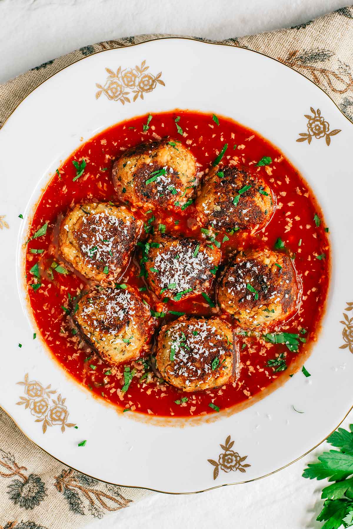 Top down view of vegan meatballs in a bowl of tomato sauce. The meatballs are nicely browned on top, big, and succulent looking. They are garnished with fresh parsley and vegan parmesan cheese.