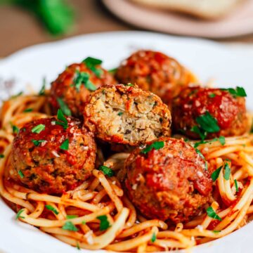 Close up of a plate of spaghetti with six vegan meatballs. The centre meatball is cut in half so the moist centre is exposed. The meatballs look large and juicy.