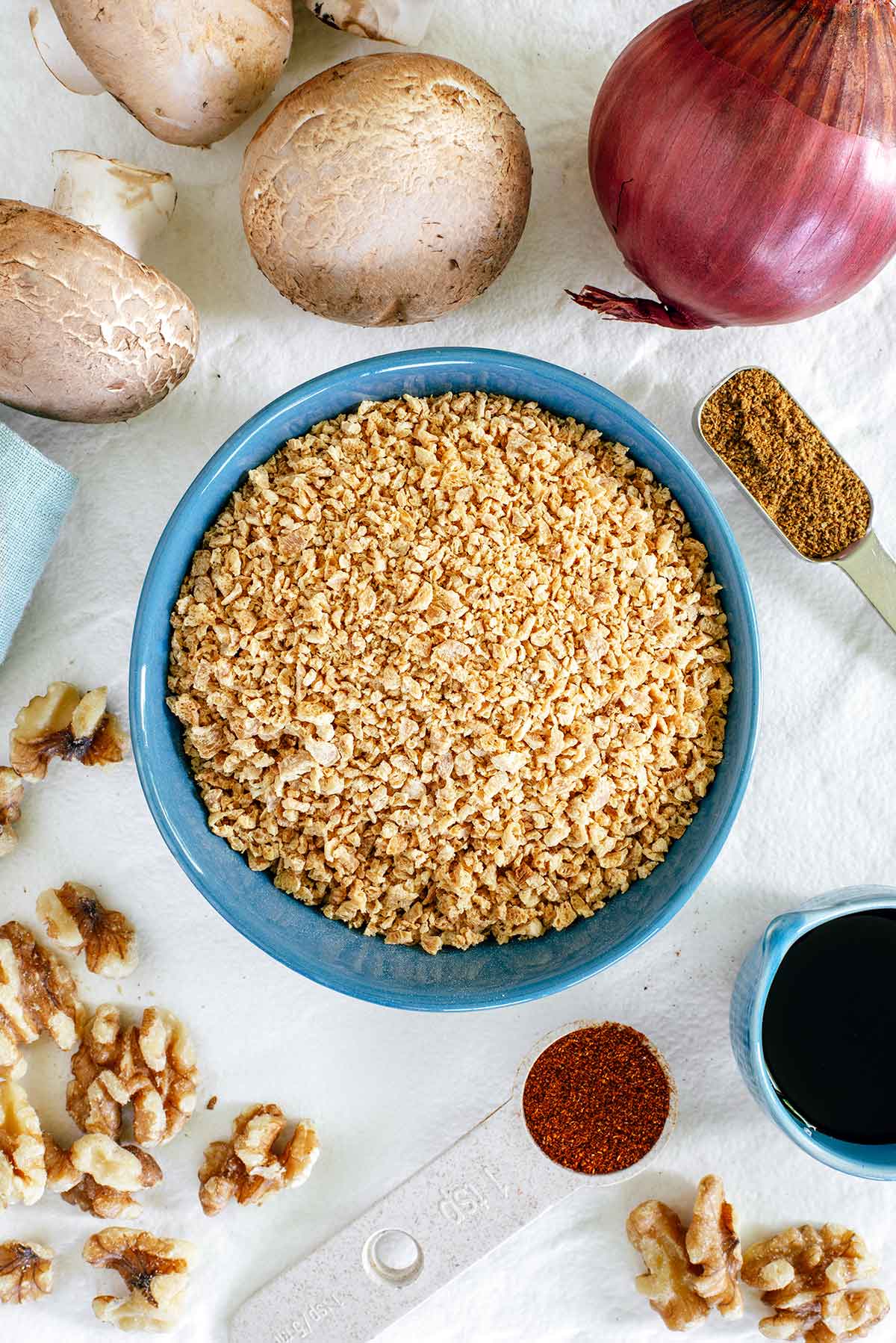 Top down view of ingredients on a white tray. A small bowl of dehydrated TVP is surrounded by cremini mushrooms, red onion, walnuts, and spices.