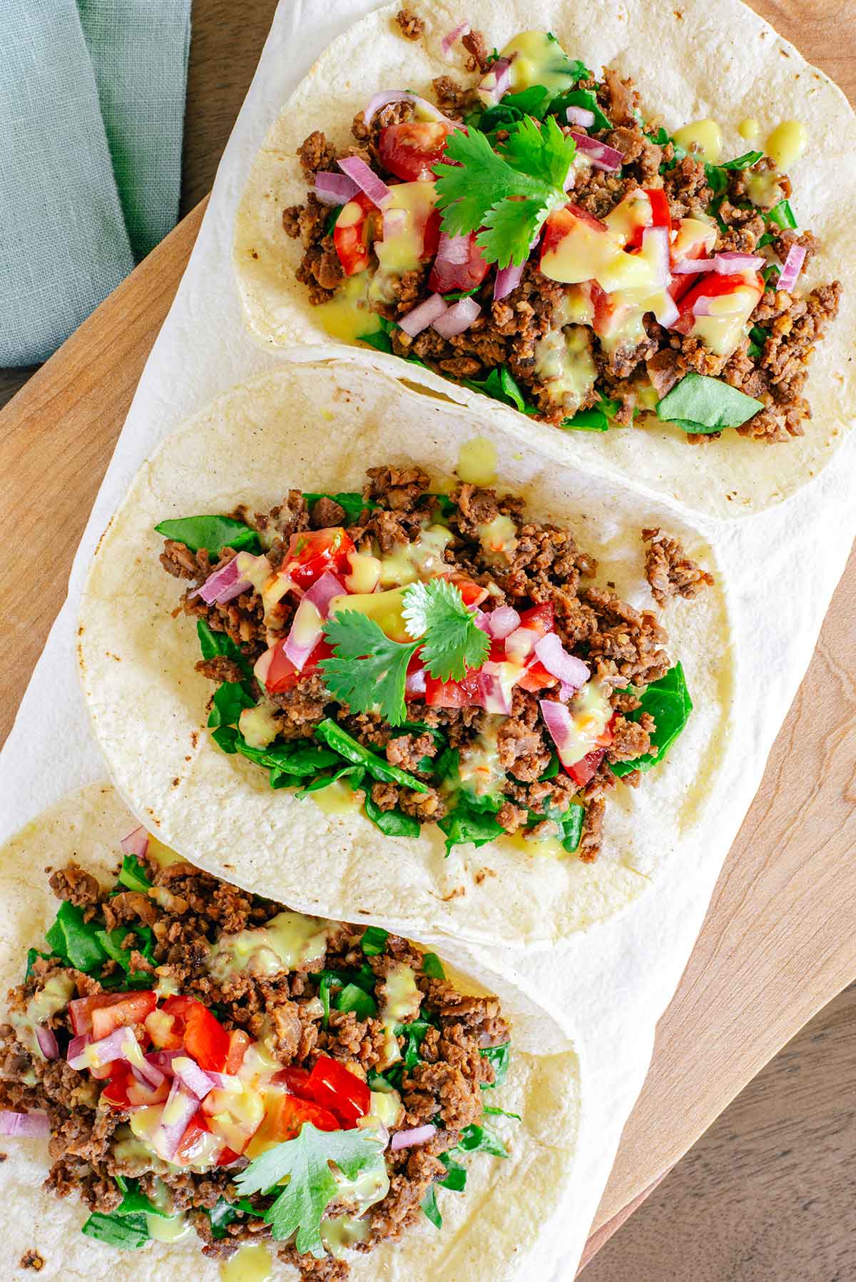 Top down view of three TVP tacos in corn tortillas dressed with chopped greens, salsa fresca, pickled red onion, vegan queso, and cilantro.