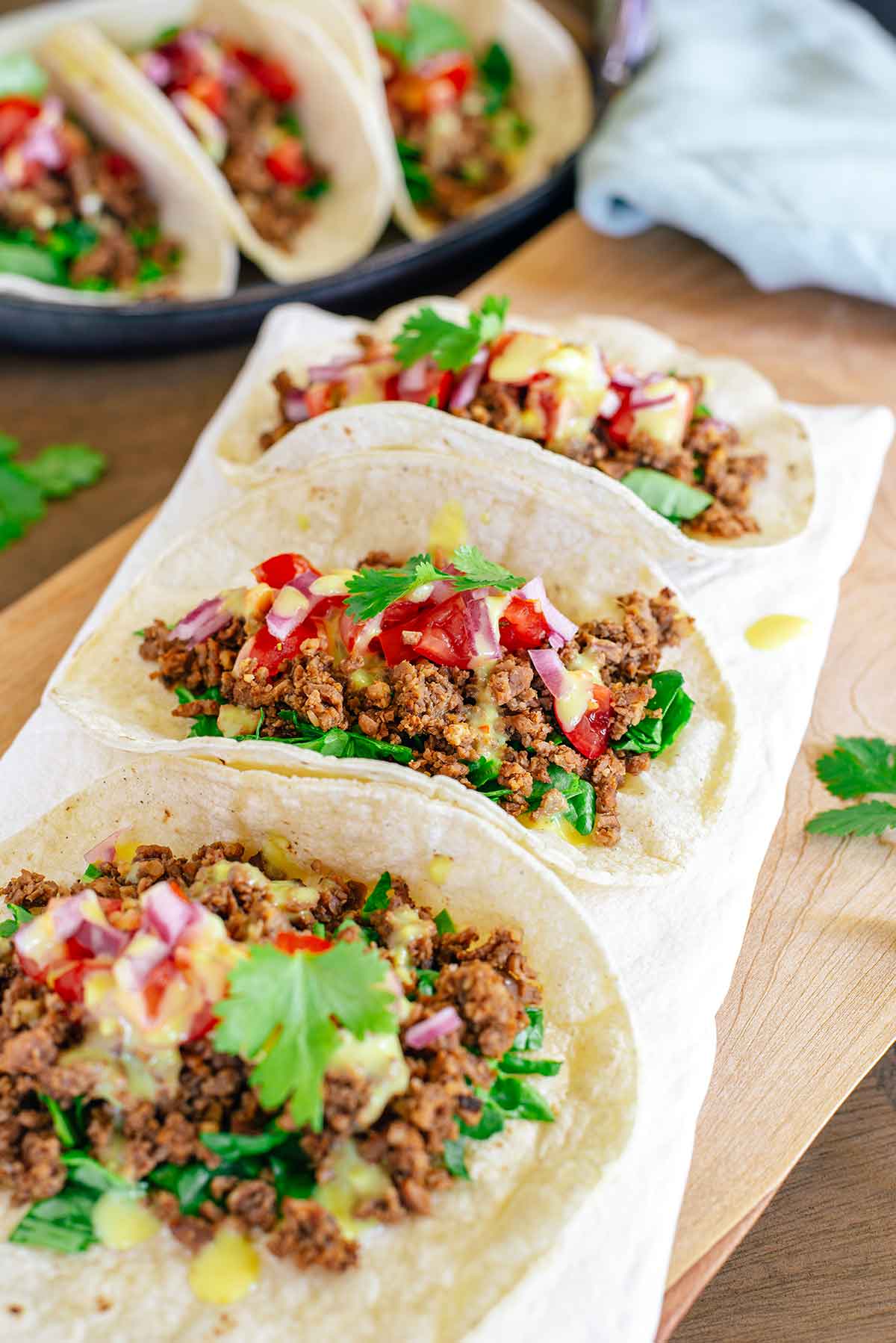 Side view of three tacos aligned on a plate. The TVP taco meat is nicely browned and crumbly but moist. The tacos are dressed with chopped greens, salsa fresca, chopped pickled red onion, vegan queso and cilantro. More tacos can be seen in the background.