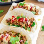 Side view of three tacos aligned on a tray. The TVP taco meat is nicely browned and crumbly but moist. The tacos are dressed with chopped greens, salsa fresca, chopped pickled red onion, vegan queso, and cilantro.