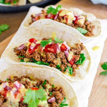 Side view of three tacos aligned on a tray. The TVP taco meat is nicely browned and crumbly but moist. The tacos are dressed with chopped greens, salsa fresca, chopped pickled red onion, vegan queso, and cilantro.