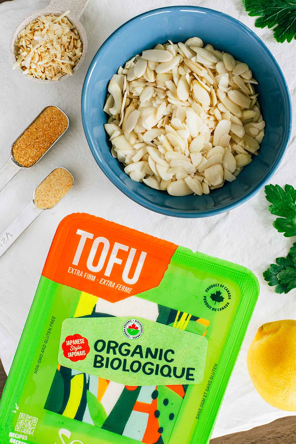 Top down view of ingredients on a white tray. A package of extra firm tofu rests beside a small bowl of sliced blanched almonds. A fresh lemon, fresh parsley, and ground seasonings fill out the tray.