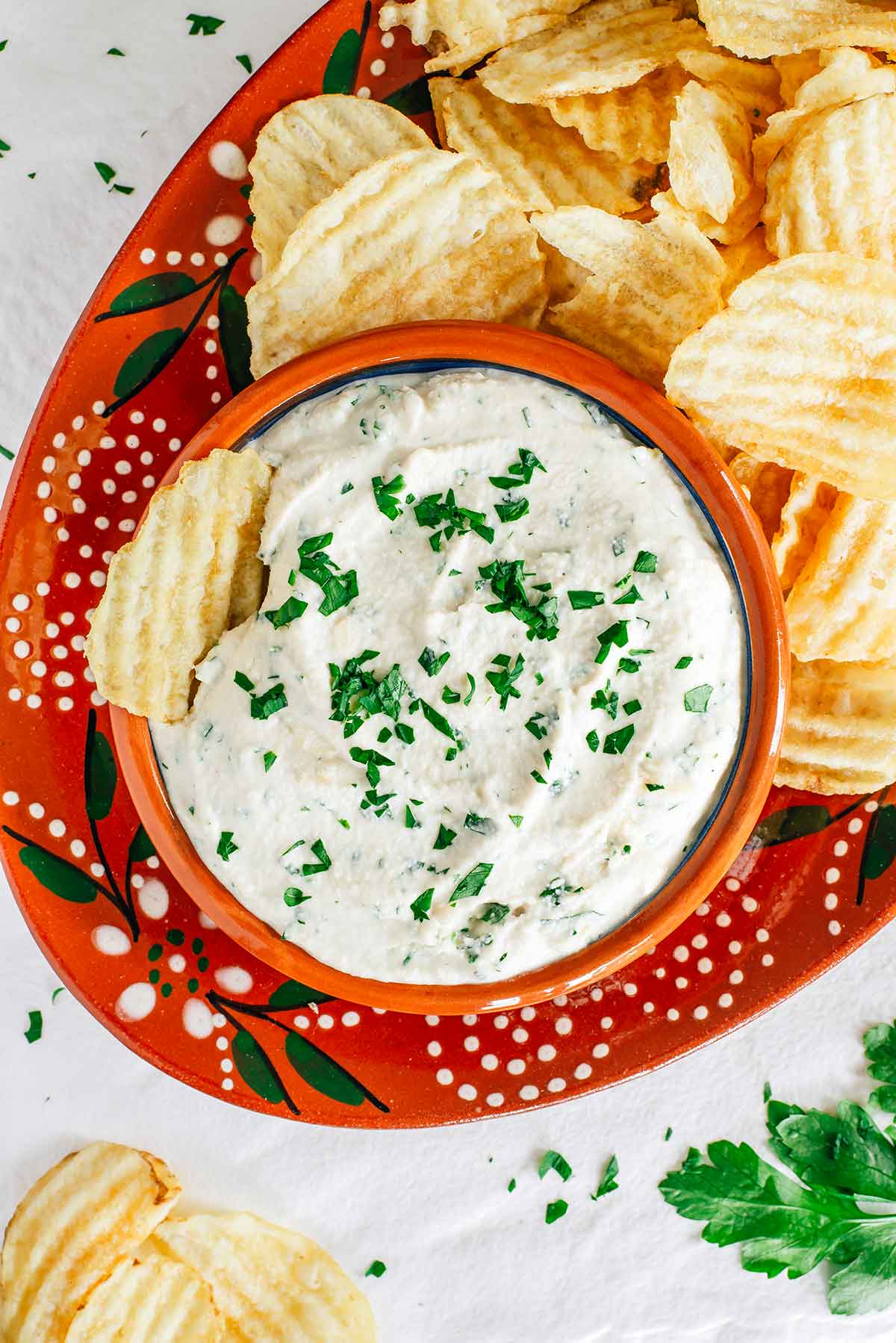 Top down view of a sour cream and onion vegan chip dip surrounded by ruffled potato chips. A chip rests in the creamy white dip which is speckled with fresh parsley.
