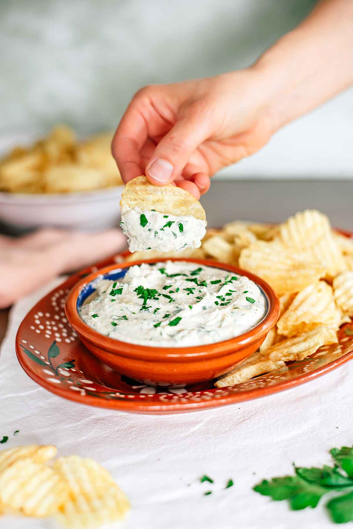 Side view of a bowl of creamy vegan chip dip surrounded by ruffled potato chips. A hand holds a chip with a dollop of dip above the bowl. The dip is creamy white and speckled with fresh chopped parsley.