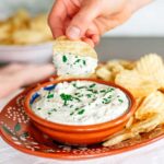 Side view of a bowl of creamy vegan chip dip. A hand holds a ruffled potato chip with a dollop of dip above the bowl. The dip is creamy white and speckled with fresh chopped parsley.