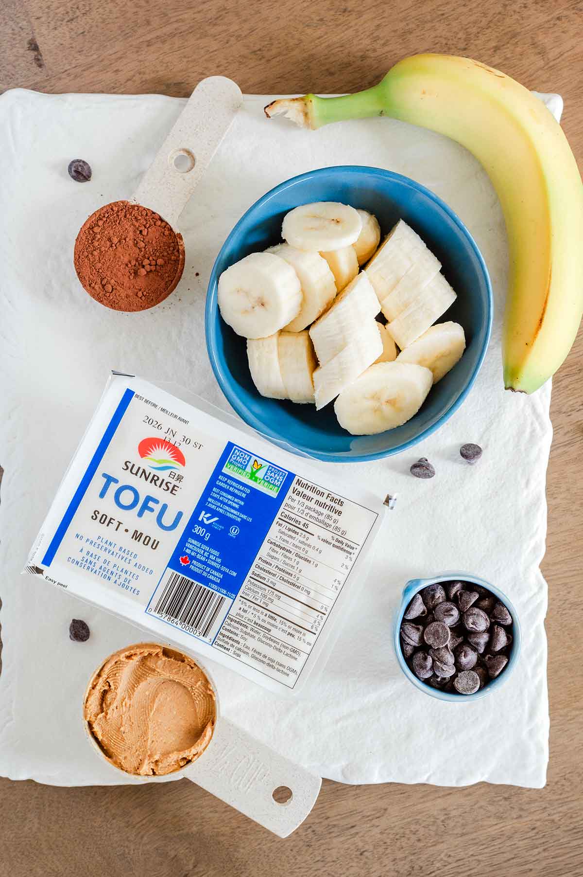 Top down view of ingredients on a white tray. A container of soft tofu is pictured next to sliced banana, peanut butter, cocoa powder, and chocolate chips.