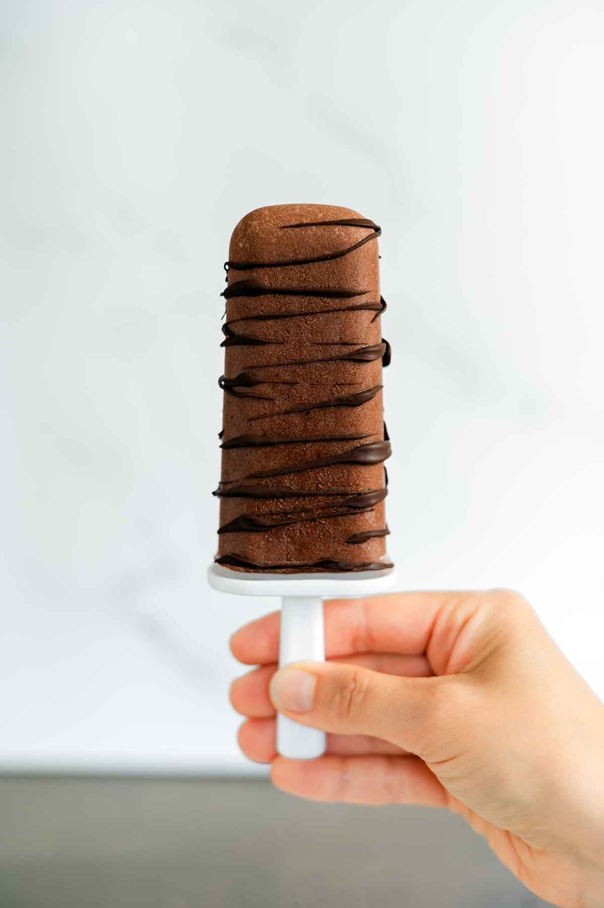 A hand holds up a vegan fudgsicle. The fudgsicle is drizzled with dark chocolate and looks like a thick and creamy frozen treat.
