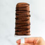 Close up of a hand holding up a vegan fudgsicle. The fudgsicle is drizzled with dark chocolate and looks like a thick and creamy frozen treat.