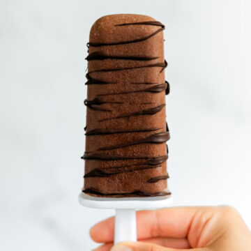 Close up of a hand holding up a vegan fudgsicle. The fudgsicle is drizzled with dark chocolate and looks like a thick and creamy frozen treat.