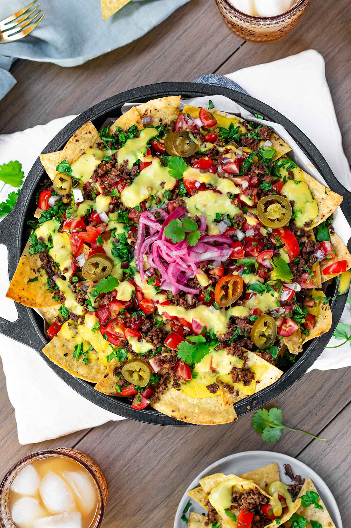 Top down view of loaded vegan nachos in a cast iron skillet on an outdoor table. Crispy tortilla chips are topped with TVP ground round, creamy vegan queso, salsa fresca, chopped greens, pickled jalapeno, pickled red onion, and fresh cilantro. 