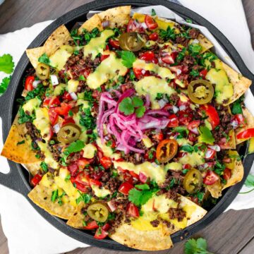 Top down view of loaded vegan nachos in a cast iron skillet. Crispy tortilla chips are topped with TVP ground round, creamy vegan queso, salsa fresca, chopped greens, pickled jalapeno, pickled red onion, and fresh cilantro.