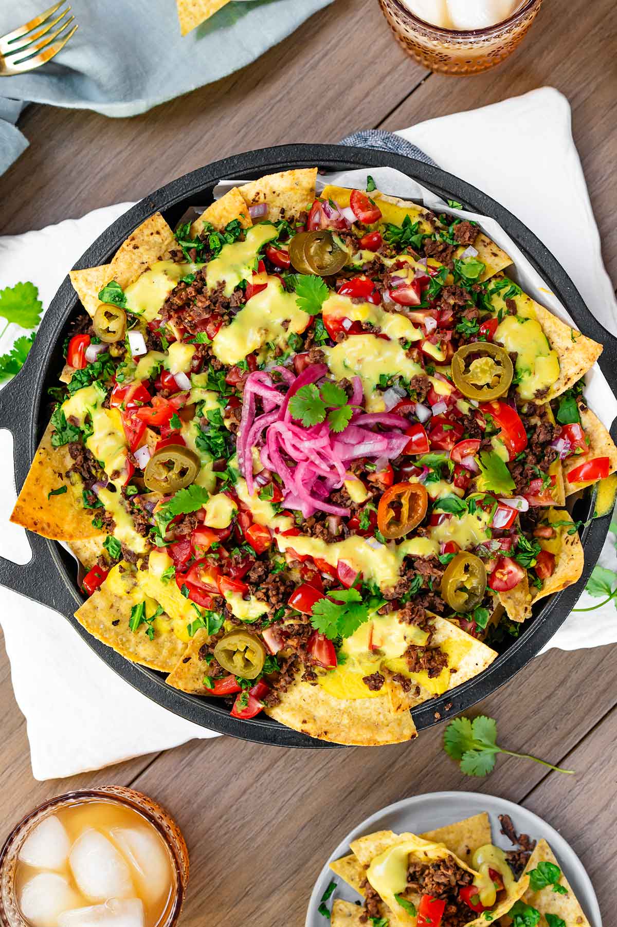 Top down view of loaded vegan nachos in a cast iron skillet on an outdoor table. Crispy tortilla chips are topped with TVP ground round, creamy vegan queso, salsa fresca, chopped greens, pickled jalapeno, pickled red onion, and fresh cilantro.