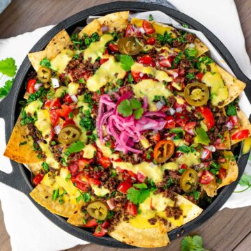 Top down view of loaded vegan nachos in a cast iron skillet. Crispy tortilla chips are topped with TVP ground round, creamy vegan queso, salsa fresca, chopped greens, pickled jalapeno, pickled red onion, and fresh cilantro.