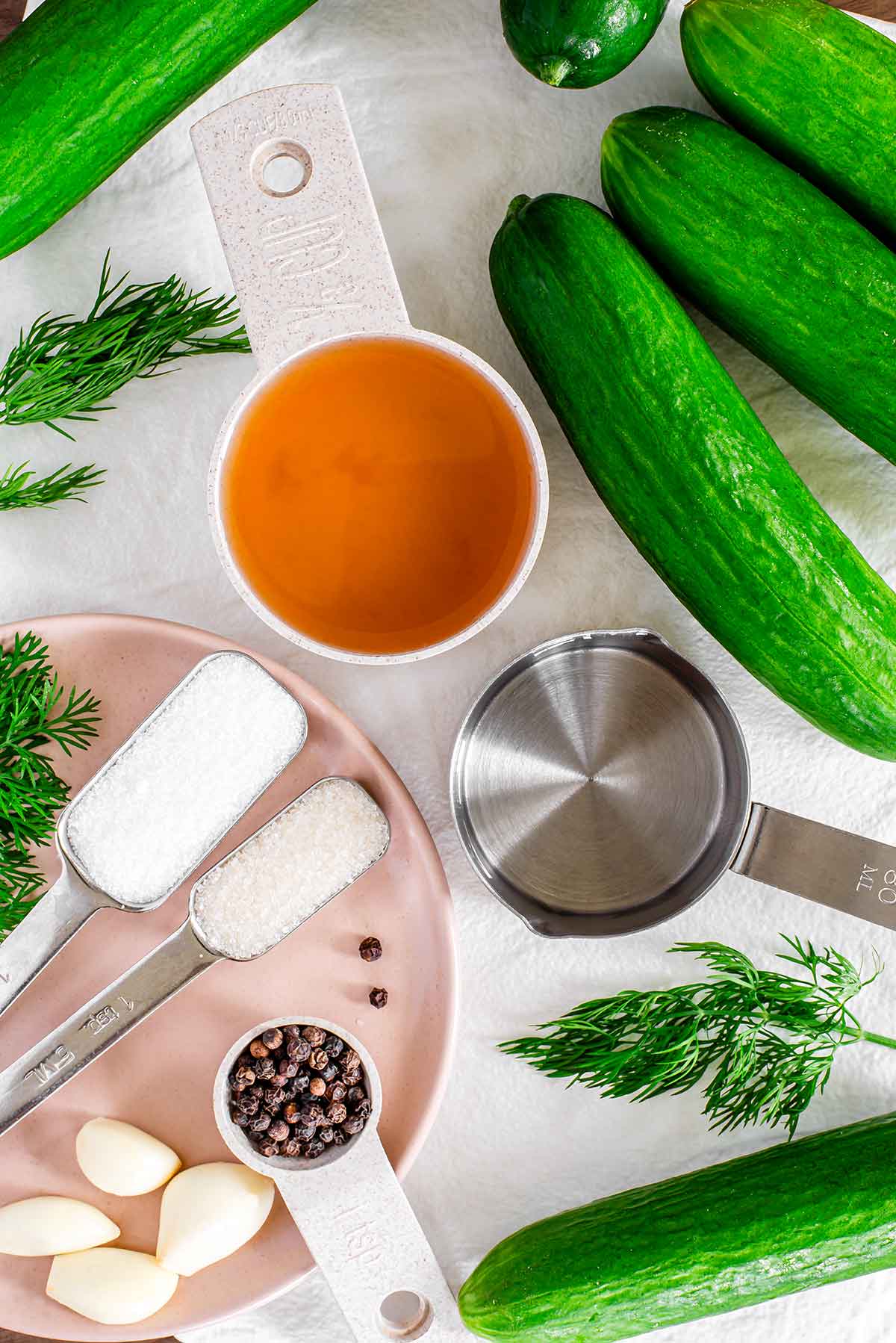 Top down view of ingredients on a white tray. Mini cucumbers are scattered around vinegar, salt, sugar, garlic cloves, black peppercorns, and fresh dill.
