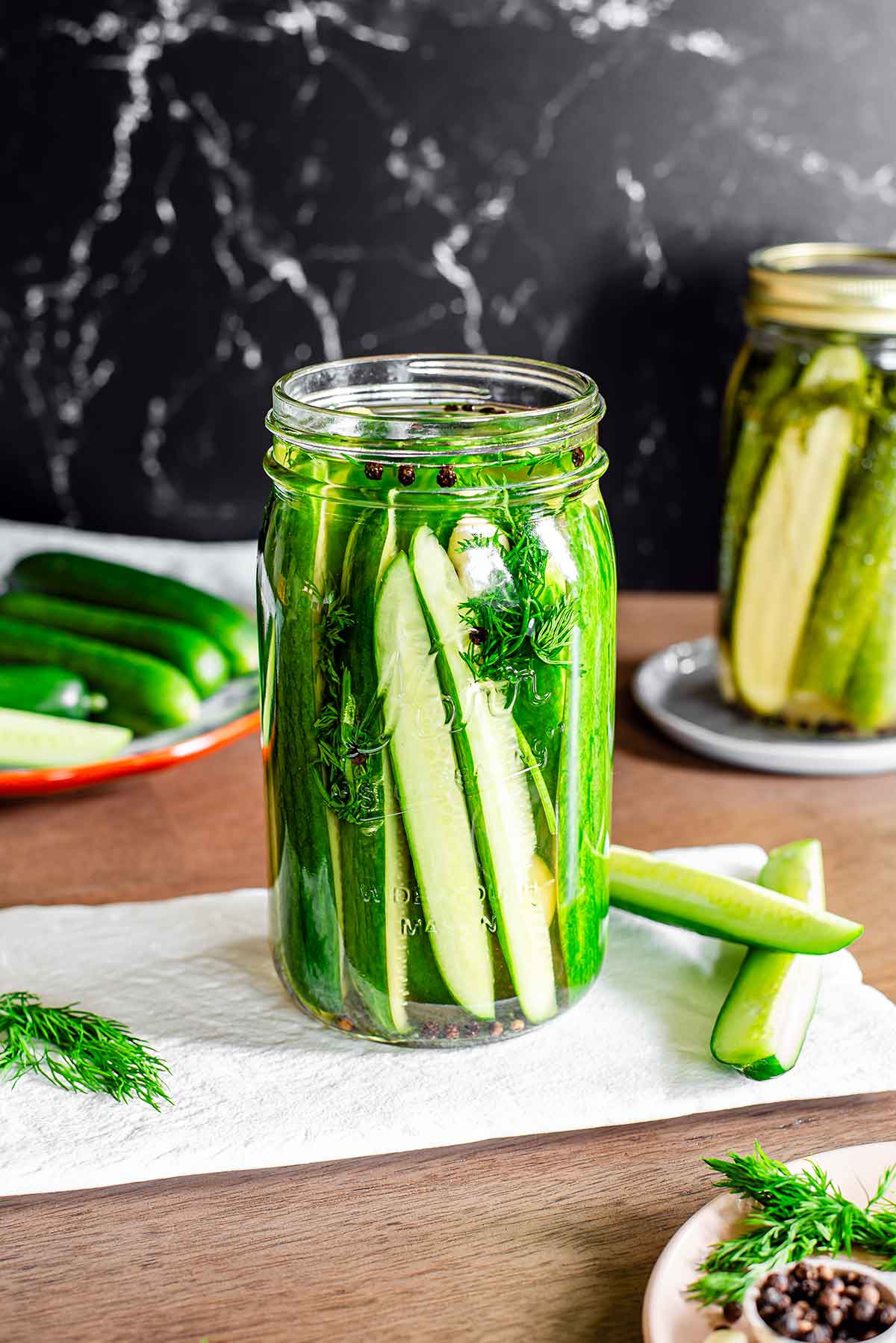 Side view of a quart jar filled with cucumber spears. The cucumber sits in a vinegar brine and is flavoured with garlic cloves, black peppercorns, and fresh dill. Another jar of older pickles is seen in the background.