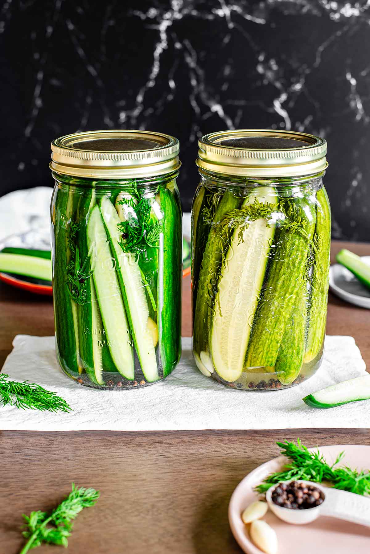 Side by side view of two 1 litre jars of refrigerator pickles. The one on the left has only just been made while the one on the right is a few weeks old. The colour of the older pickles has become less vibrant. Both are flavoured with garlic cloves, peppercorns, and fresh dill.