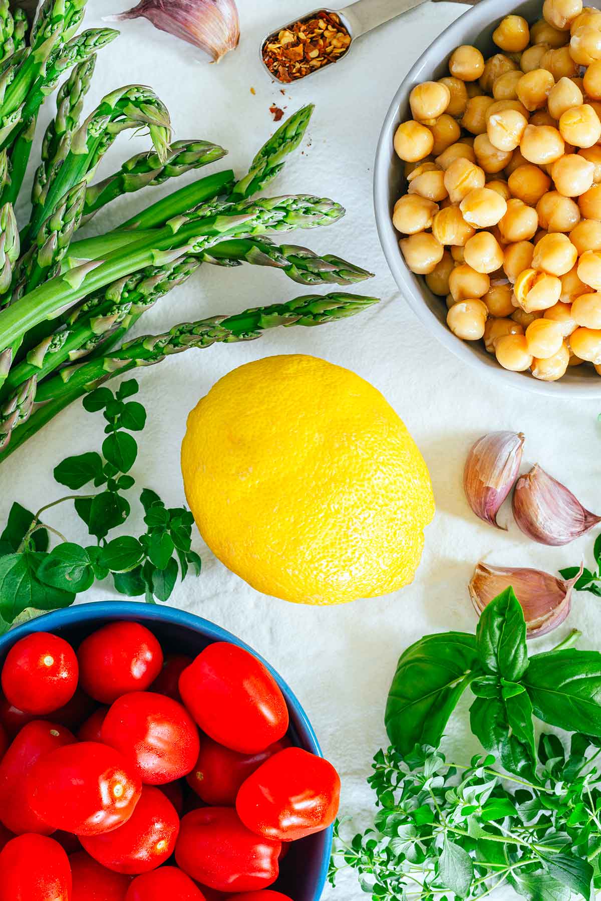 Top down view of ingredients on a white tray. A large lemon sits in the middle of asparagus spears, grape tomatoes, fresh herbs, garlic cloves, chickpeas, and red chili flakes. 
