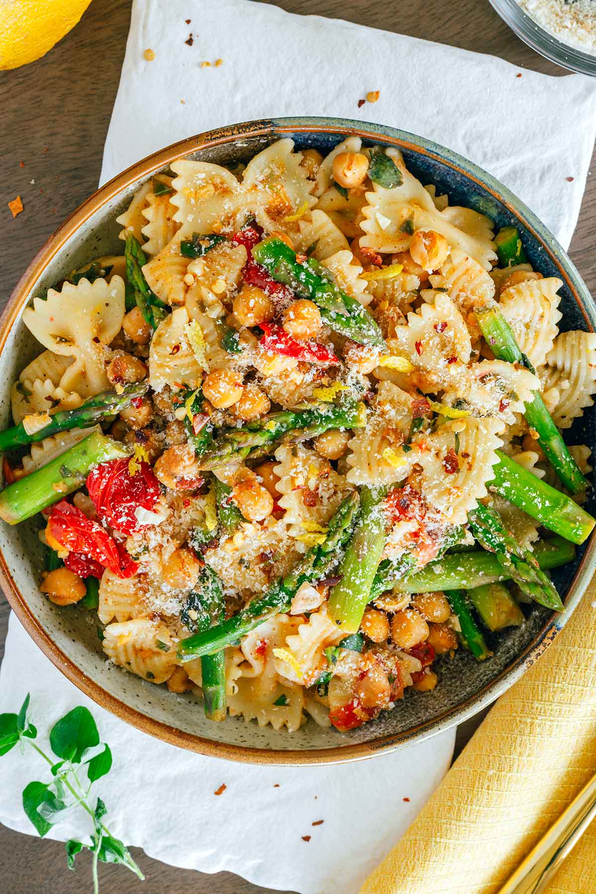 Top down view of a bow tie pasta dish with asparagus, grape tomatoes and chickpeas. A light clear lemony dressing with chili flakes, garlic, and herbs, coats the dish. A fresh lemon and fresh oregano can be seen on the table around the bowl of pasta.  