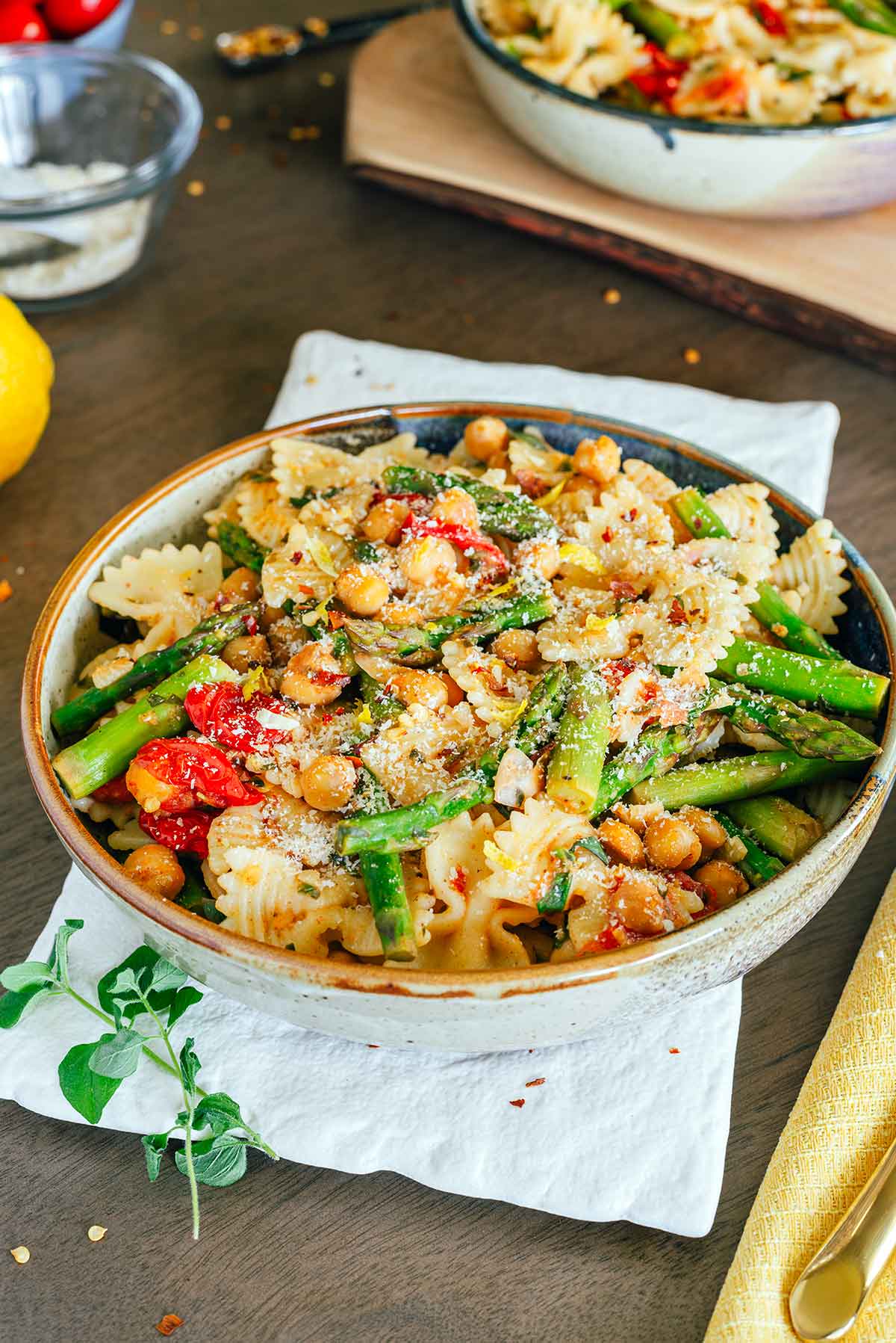 Side view of a colourful bowl of lemon asparagus pasta. Bow tie pasta is mixed with green asparagus spears, red grape tomatoes, and chickpeas. Fresh green herbs, red chili flakes, and lemom zest are speckled across the pasta. 