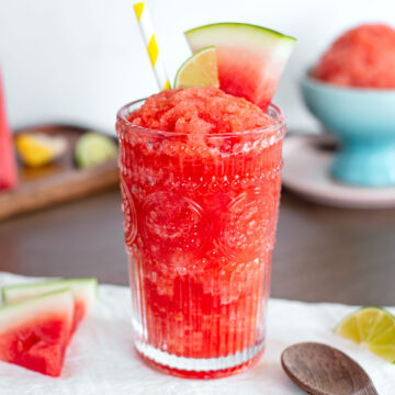 Close up side view of a pink watermelon slushie in a decorative glass. The frosty drink is garnished with small slices of watermelon and lime. A colourful striped straw rests in the thick slushie.