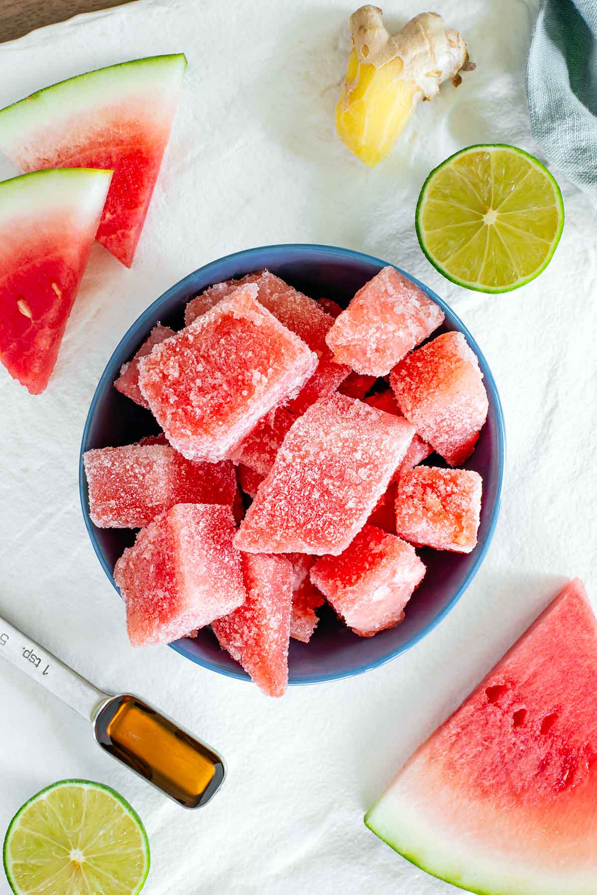 Top down view of cubed frozen watermelon in a small dish on a white tray. Slices of fresh watermelon, a halved lime, a teaspoon of maple syrup, and a knob of ginger surround.