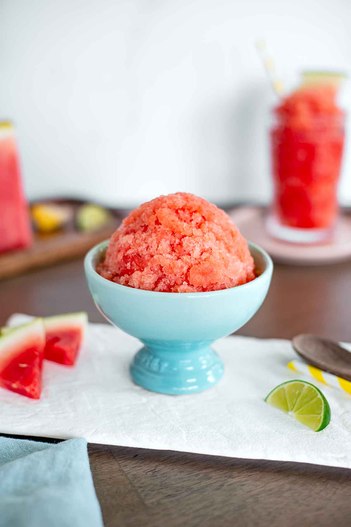 Side view of a pink watermelon snow cone. The icy treat is shaped into a dome in a ceramic ice cream cup. Fresh watermelon, a slice of lime, a colourful straw, and a spoon rest beside the snow cone.