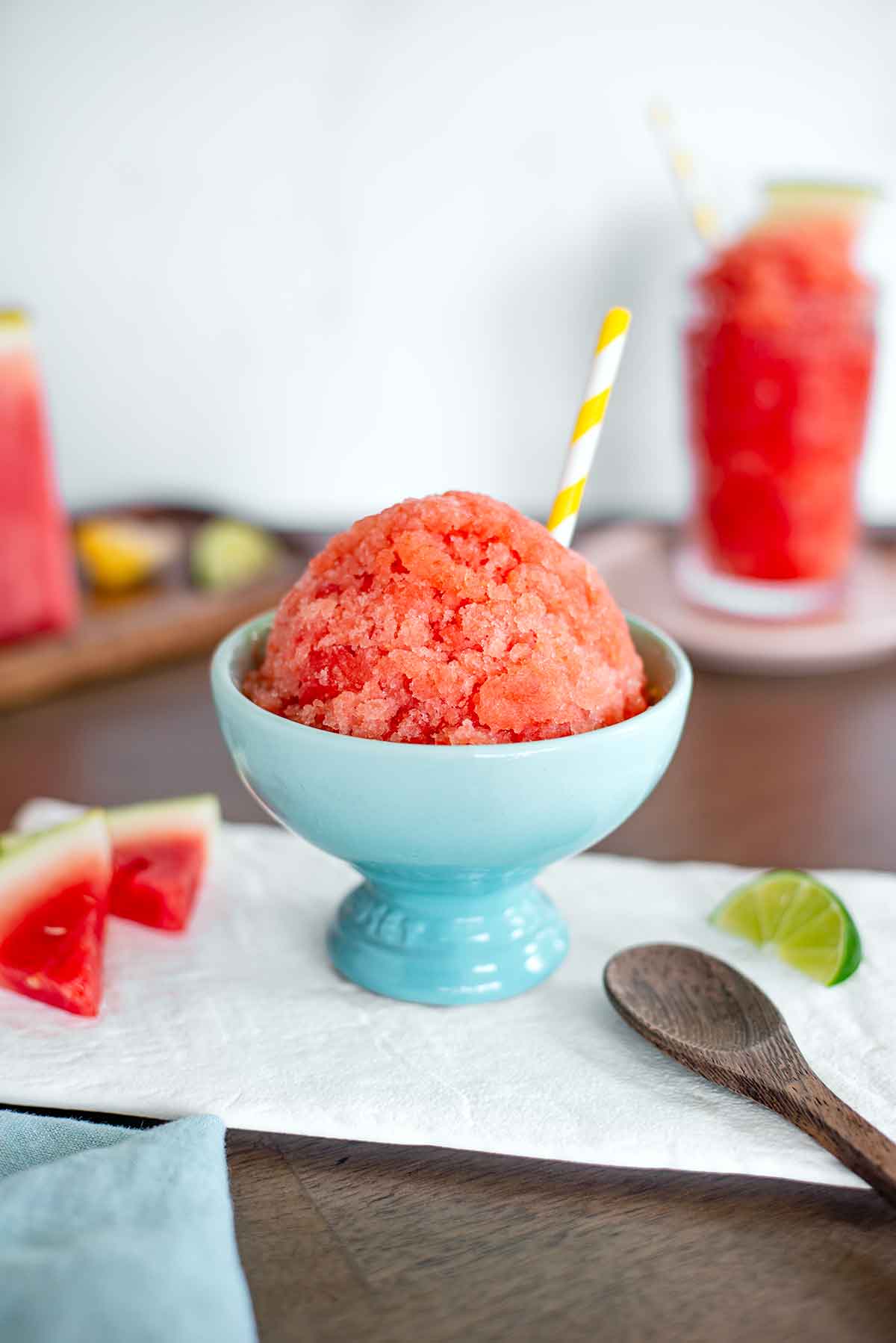 Side view of a domed watermelon snow cone in an ice cream cup. The frozen watermelon treat is crystallized and bright pink. A yellow straw pokes out from the snow cone and a spoon rests beside the bowl. A tall watermelon slushie drink can be seen in the background.