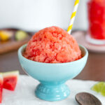 Side view of a domed watermelon snow cone in an ice cream cup. The frozen watermelon treat is crystallized and bright pink. A yellow straw pokes out from the snow cone and a spoon rests beside the bowl.