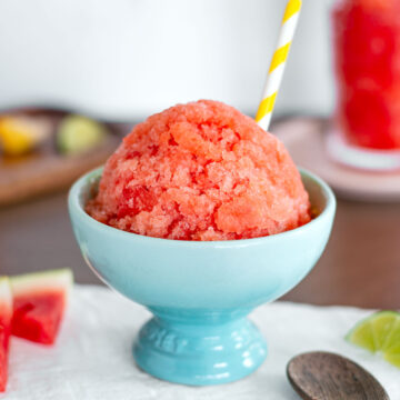 Side view of a domed watermelon snow cone in an ice cream cup. The frozen watermelon treat is crystallized and bright pink. A yellow straw pokes out from the snow cone and a spoon rests beside the bowl.