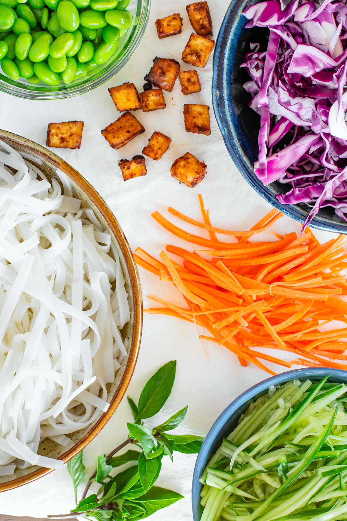 Top down view of ingredients on a white tray. Rice noodles, crispy baked tofu cubes, shelled edamame, shredded red cabbage, julienned carrot and cucumber, and Thai basil leaves are assembled.