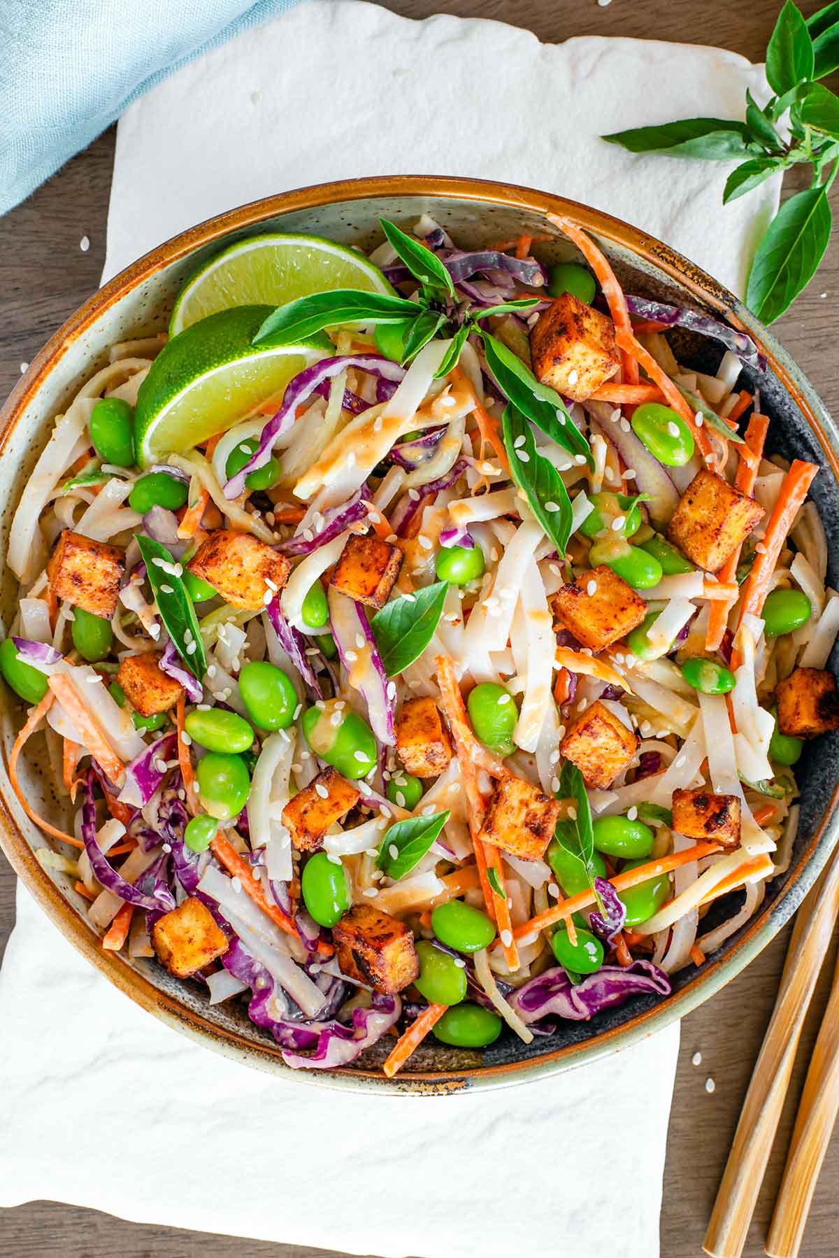 Top down view of a colourful cold noodle salad with rice noodles, red cabbage, edamame, carrot and cucumber. The salad is tossed in a peanut sauce and topped with crispy tofu. Lime wedges and Thai basil leaves garnish. Chopsticks rest beside the beautiful salad bowl.