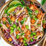 Top down view of a colourful cold noodle salad with rice noodles, red cabbage, edamame, carrot and cucumber. The salad is tossed in a peanut sauce and topped with crispy tofu. Lime wedges and Thai basil leaves garnish.