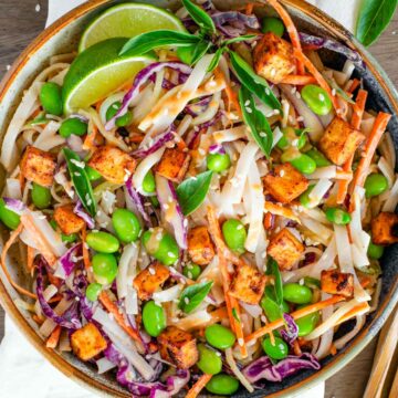 Top down view of a colourful cold noodle salad with rice noodles, red cabbage, edamame, carrot and cucumber. The salad is tossed in a peanut sauce and topped with crispy tofu. Lime wedges and Thai basil leaves garnish.